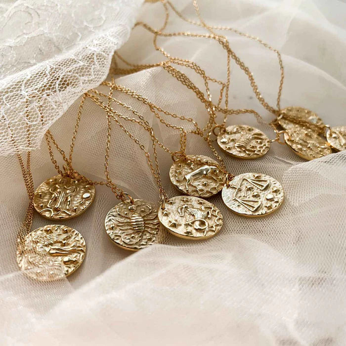 Collection de colliers astrologiques en plaqué or 18 carats, pendentifs ronds gravés des symboles des signes du zodiaque, chaßnes fines élégantes, présentation sur tissu blanc léger. Idée cadeau femme.