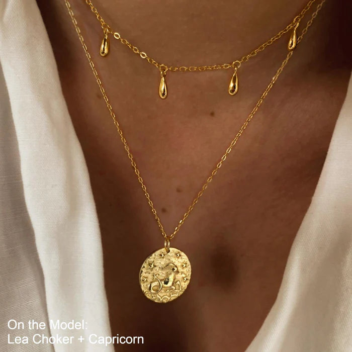 Collier doré avec pendentif signe Capricorne porté avec un autre collier à pampilles, sur une peau hùlée et un chemisier blanc. Idée cadeau femme.