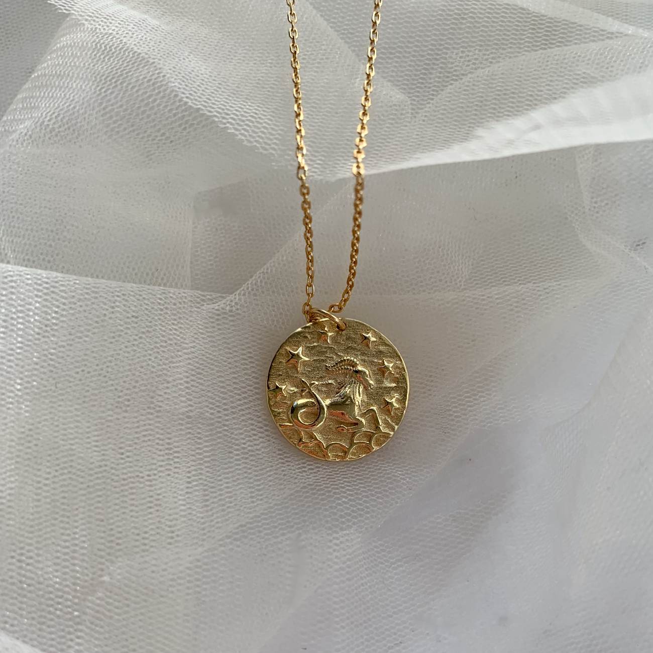 Collier dorĂ© signe astrologique Capricorne posĂ© sur voile blanc, pendentif gravĂ© dâune chĂšvre et dâĂ©toiles. IdĂ©e cadeau femme.