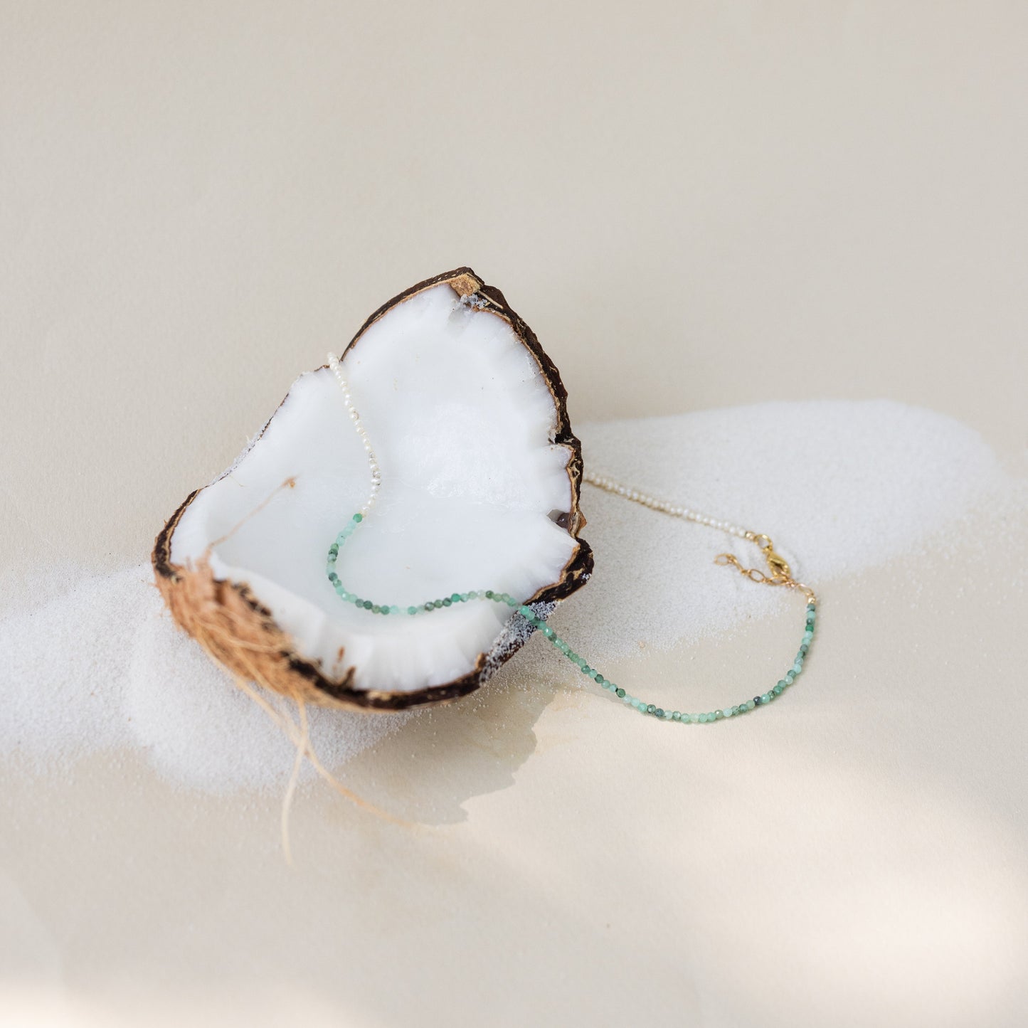Collier Emmie en perles de culture et pierres émeraude, bijou artisanal délicatement posé sur une moitié de noix de coco sur sable fin, idée cadeau femme