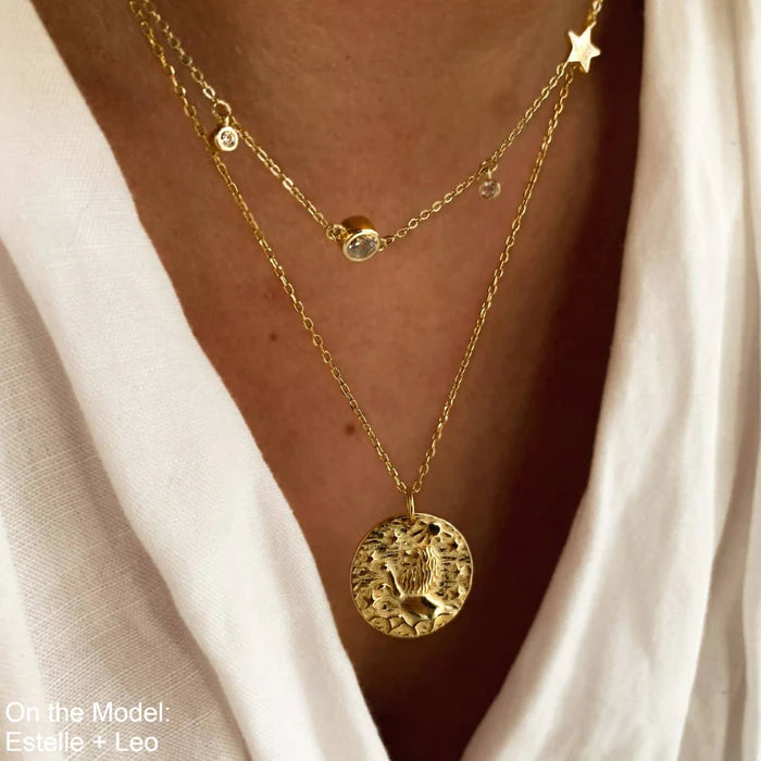 Collier en plaquĂ© or 18 carats avec pendentif rond illustrant un lion stylisĂ© entourĂ© dâĂ©toiles, portĂ© par une femme en chemise blanche. IdĂ©e cadeau femme.