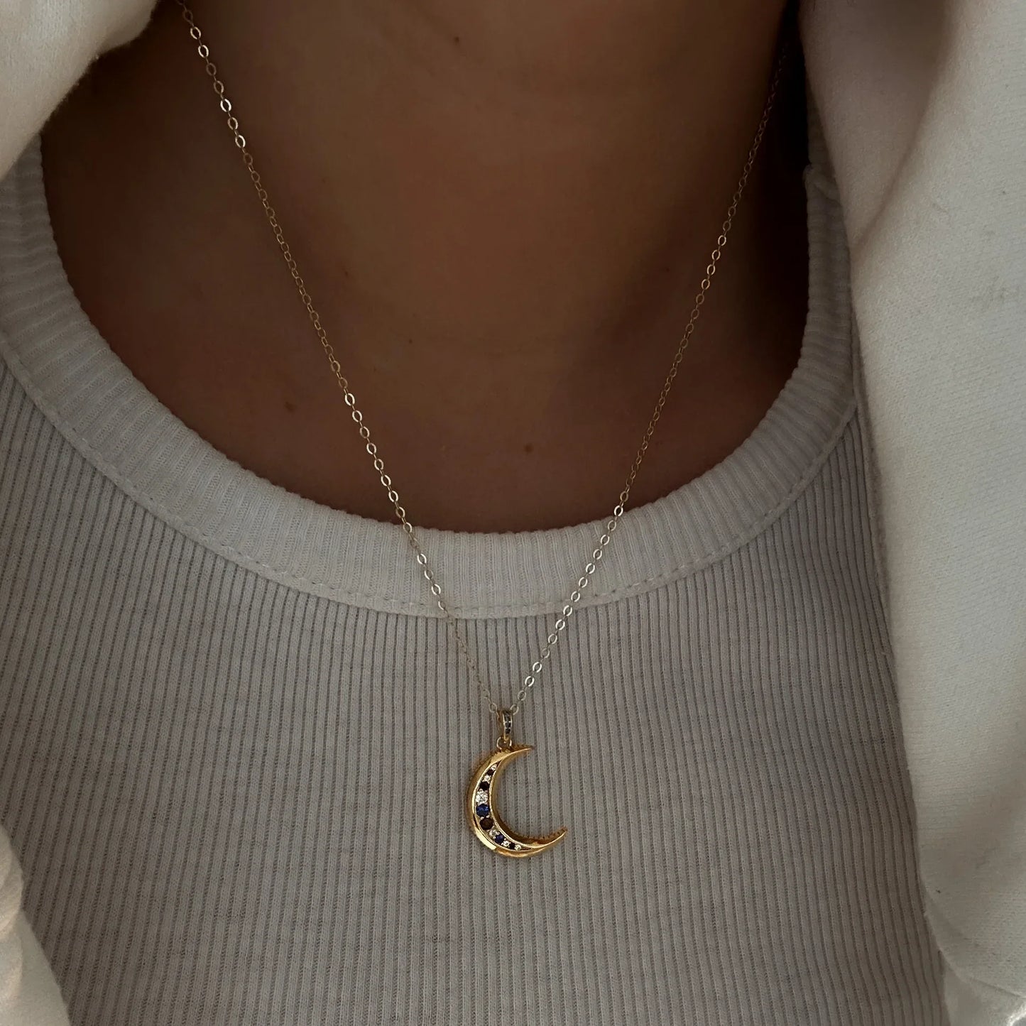 Femme portant un collier lune doré avec pierres colorées sur un pull clair. Idée cadeau femme.