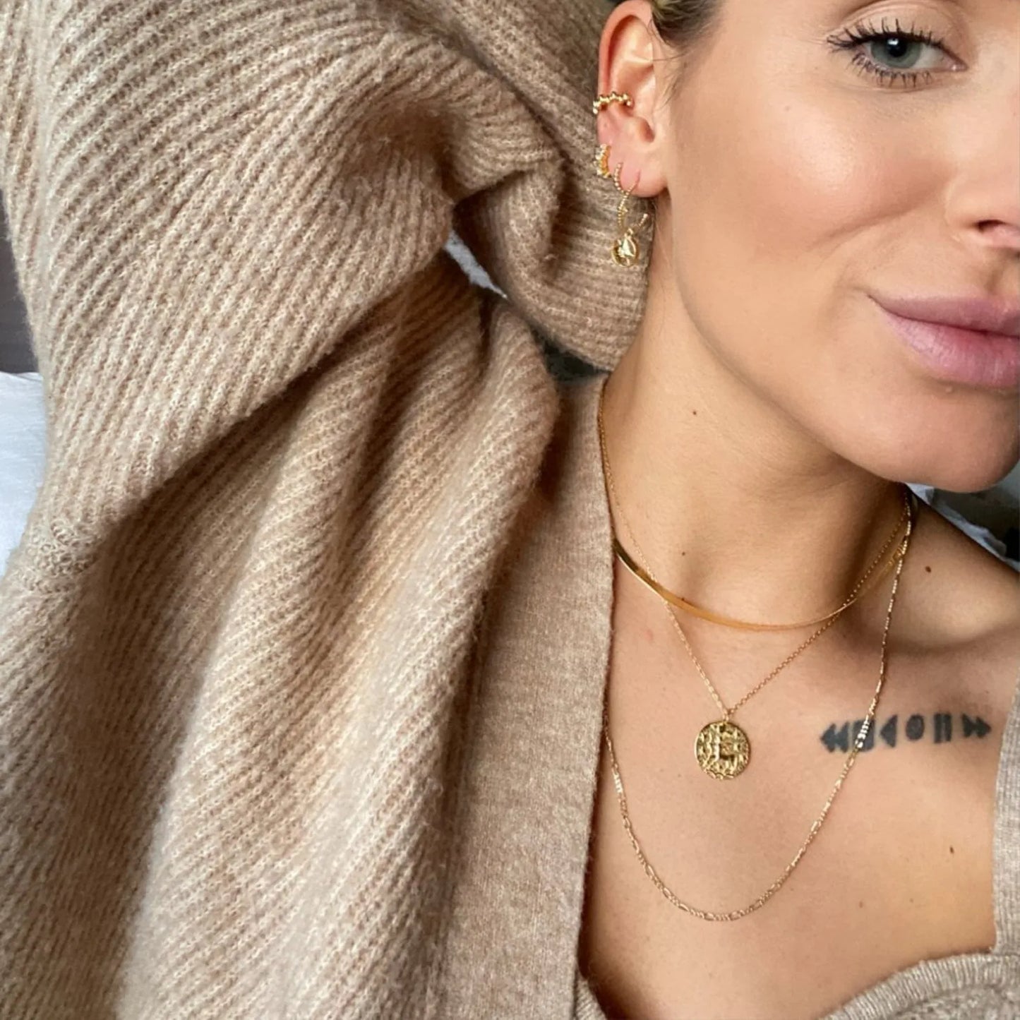 Collier sagittaire en plaquĂ© or 18 carats portĂ© en double chaĂźne par une femme, pendentif rond gravĂ© du symbole Balance entourĂ© dâĂ©toiles, look tendance avec superposition de bijoux, portĂ© avec pull beige. IdĂ©e cadeau femme.