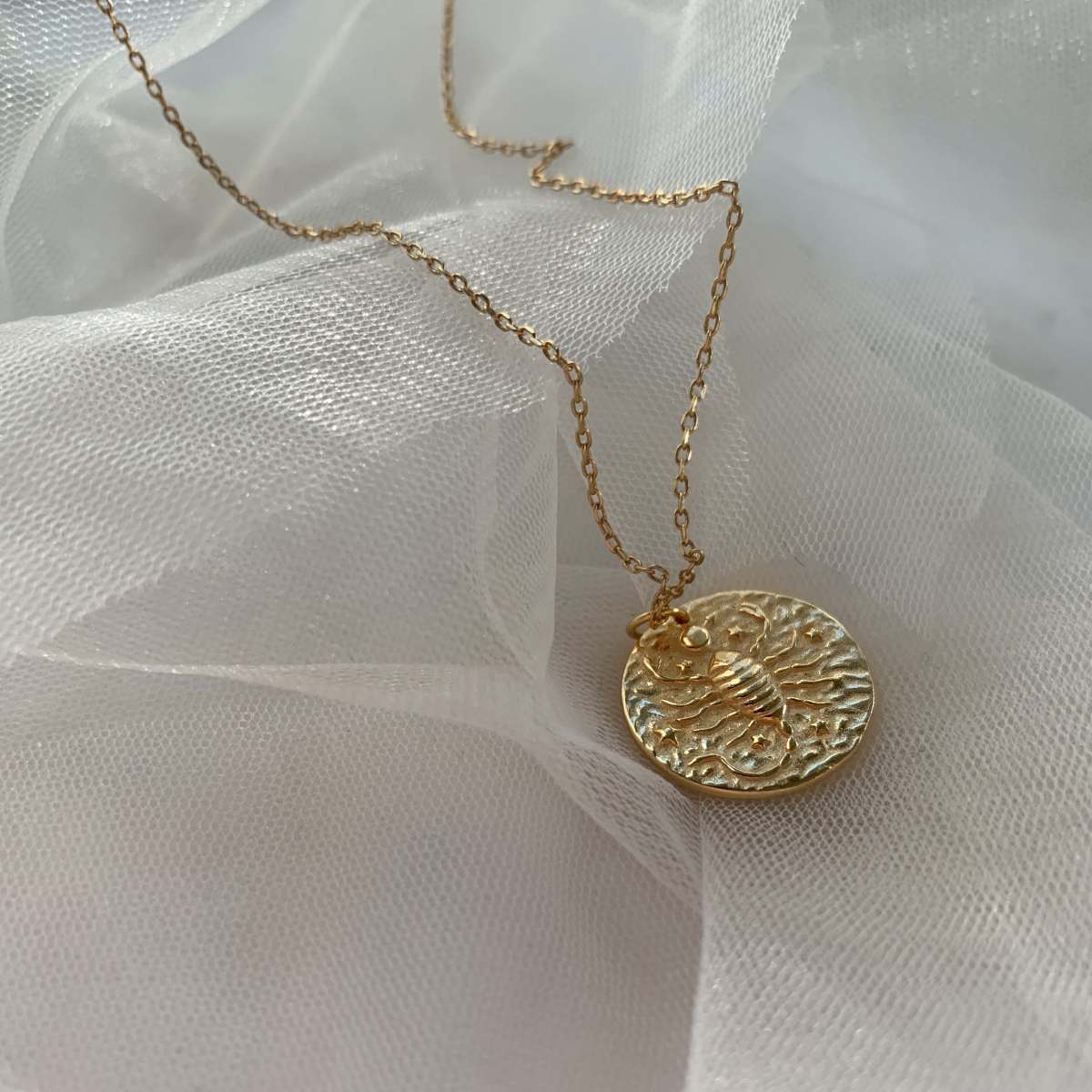 Collier Scorpion en plaquĂ© or 18 carats avec pendentif rond texturĂ© gravĂ© dâun scorpion entourĂ© dâĂ©toiles, chaĂźne fine dĂ©licate, bijou fĂ©minin Ă©lĂ©gant posĂ© sur tissu blanc lĂ©ger. IdĂ©e cadeau femme.