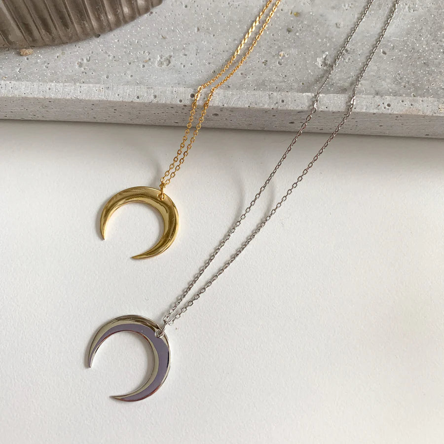 ALT TEXT : Deux colliers croissant de lune en version dorée et argentée, posés côte à côte sur fond clair.
