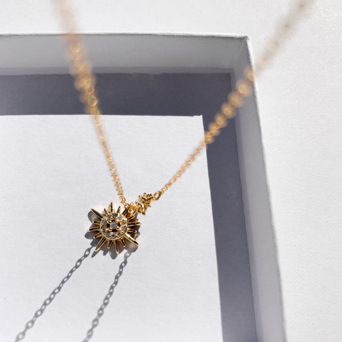 Idées cadeaux pour femme : collier double pendentif soleil présenté dans sa boîte cadeau, capturant la lumière pour un effet poétique et raffiné
