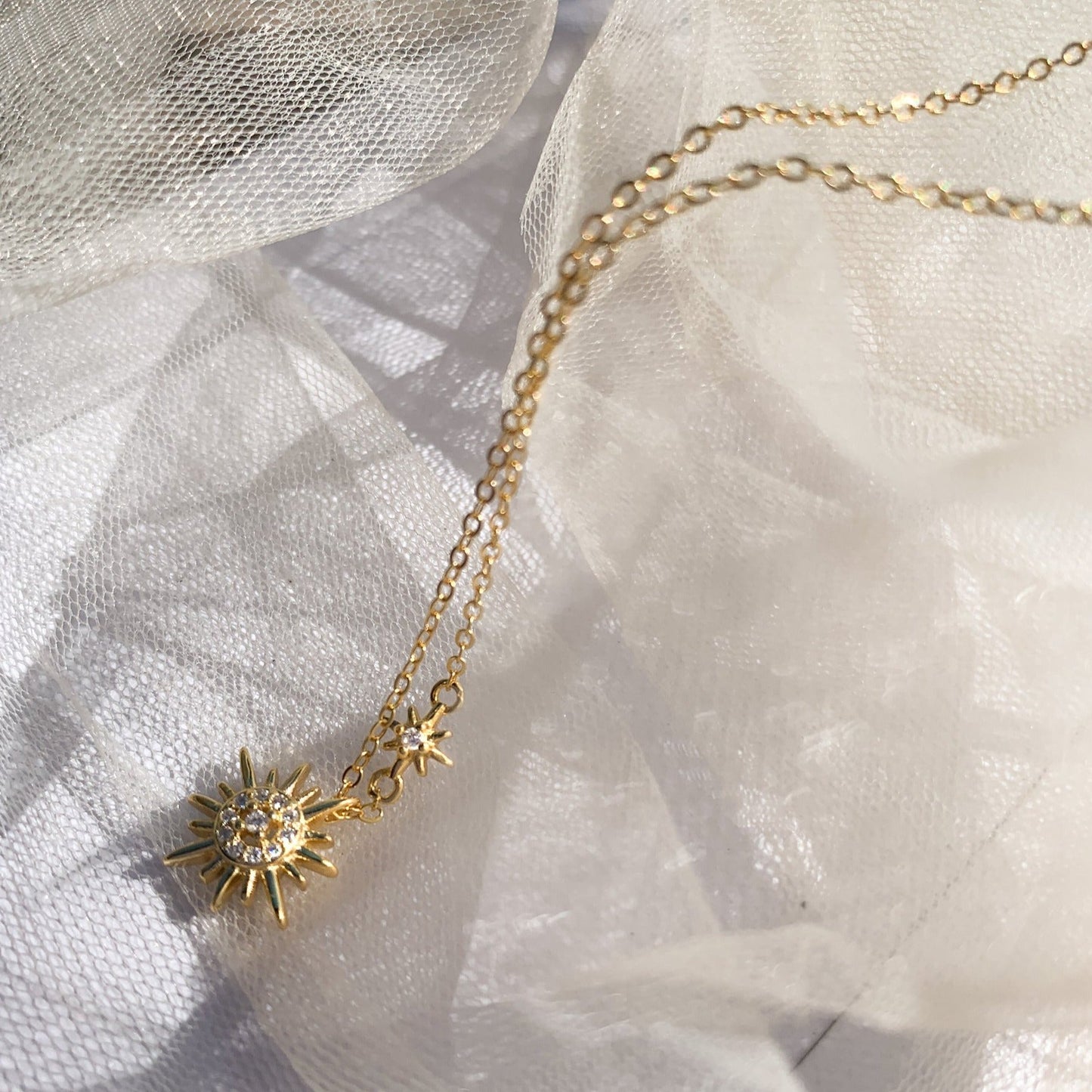 Idées cadeaux pour femme : collier double pendentif soleil en zircons, mis en scène sur fond de tulle, pour un rendu délicat, lumineux et poétique