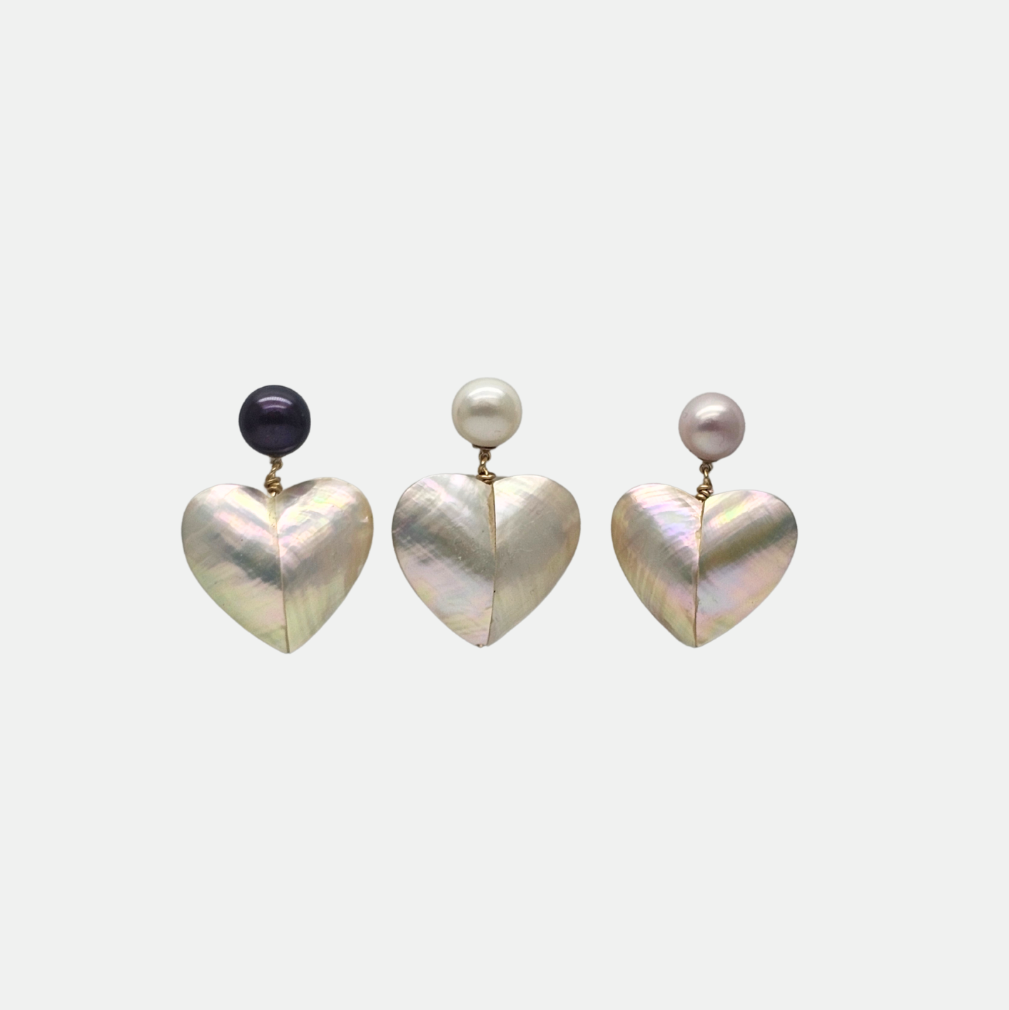 Gros plan sur trois boucles d'oreilles pendantes en forme de cœur en nacre blanche irisée, chacune surmontée d'une perle de culture baroque ronde dans des couleurs différentes : noire, crème et rose pâle. Les montures sont en plaqué or. Le bijou est élégant et idéal comme cadeau. koolkado, cadeau.