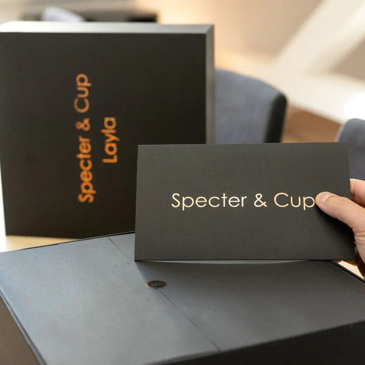 Main tenant la carte de bienvenue Specter et Cup devant le coffret de luxe Layla illustrant un emballage haut de gamme exclusif