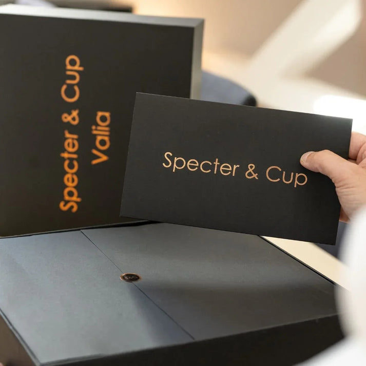 Main tenant la carte de bienvenue Specter et Cup devant le coffret de luxe Valia illustrant un emballage haut de gamme exclusif pour offrir