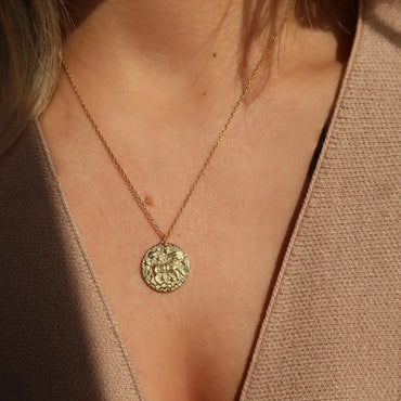  Collier doré avec pendentif Bélier porté sur une femme en top beige, éclat doré mis en valeur par la lumière naturelle. Idée cadeau femme.
