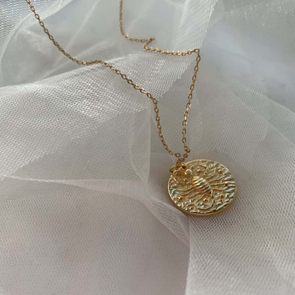 Collier Scorpion en plaqué or 18 carats avec pendentif rond texturé gravé d’un scorpion entouré d’étoiles, chaîne fine délicate, bijou féminin élégant posé sur tissu blanc léger. Idée cadeau femme.