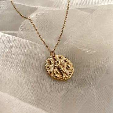 Collier Verseau en plaqué or 18 carats avec pendentif rond texturé gravé d’un verseau entouré d’étoiles et planètes, bijou féminin raffiné posé sur tissu blanc léger. Idée cadeau femme.